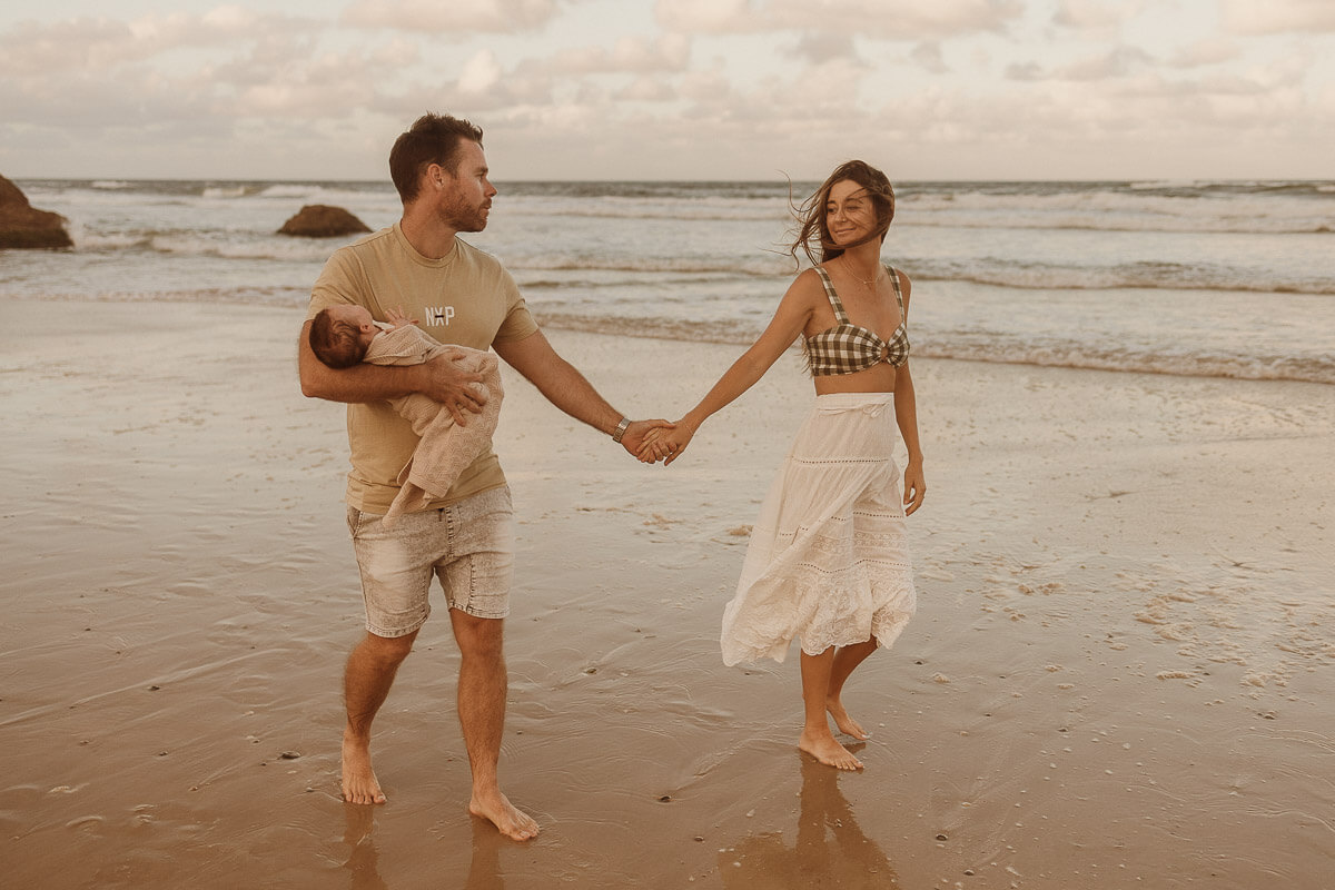 Couple with newborn wander by the water's edge at the beach for their newborn session with Photos by Jordi.