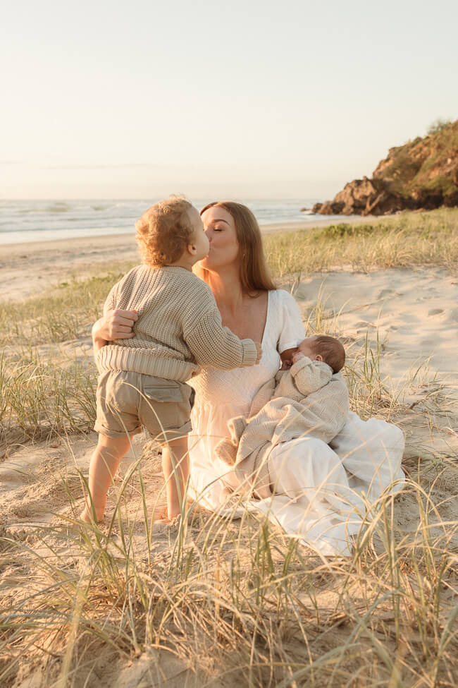 A mother shares a sweet moment with her sons during a newborn photo session on the beach at sunrise.