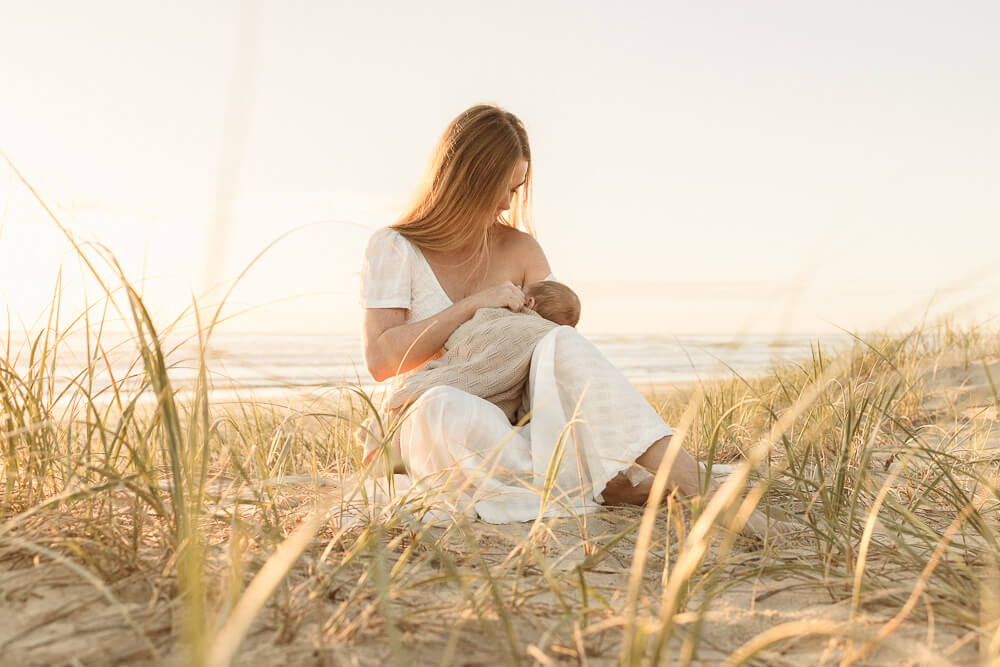 A mother breastfeeds her newborn son amongst the dunes at sunrise.