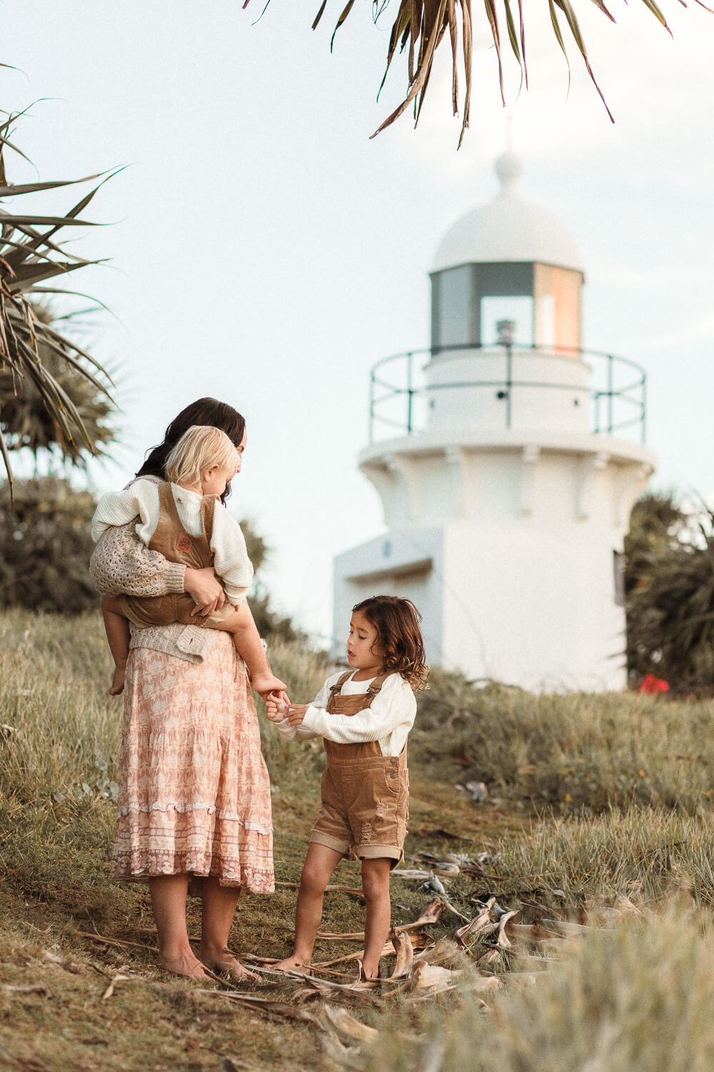 What-to-wear-to-a-family-photo-session-18 A person stands on a grassy path near a white lighthouse, holding a small child and looking at an older child beside them. The older child is holding the hand of the adult. Palm trees frame the scene. The sky is lightly clouded, indicating either early morning or late afternoon—captured by a motherhood photographer Gold Coast.