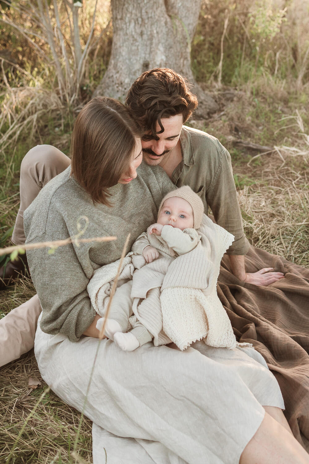 Winter-photoshoot-Gold-Coast-01 Young couple sitting under a tree during a winter photoshoot, lovingly holding their baby wrapped in soft, neutral layers on the Gold Coast.