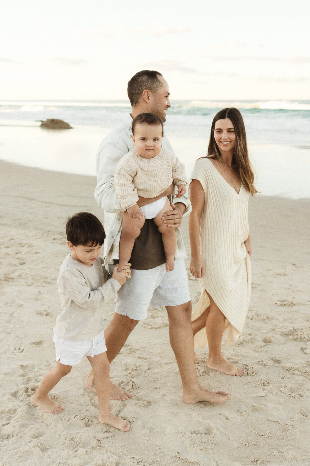 Winter-photoshoot-Gold-Coast-02 Young family walking barefoot along the beach during a winter photoshoot on the Gold Coast, dressed in soft neutral tones with the ocean in the background.