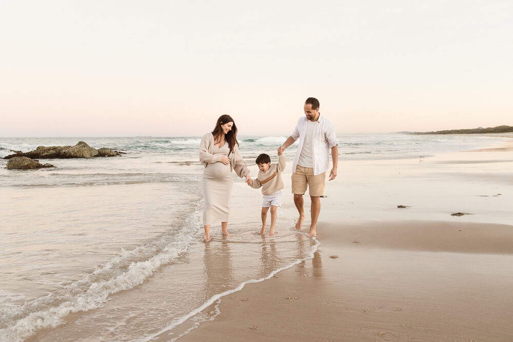 Winter-photoshoot-Gold-Coast-06 Pregnant mother, father, and young child walking barefoot along the shoreline during a winter photoshoot on the Gold Coast, with soft pastel skies and gentle waves.