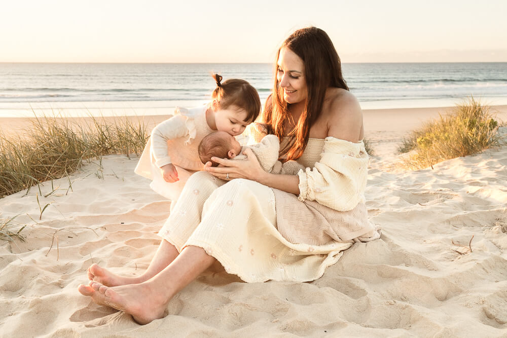 Winter-photoshoot-Gold-Coast-08 Mother sitting on the sand at the beach during a winter photoshoot on the Gold Coast, holding her newborn while her toddler gently kisses the baby.